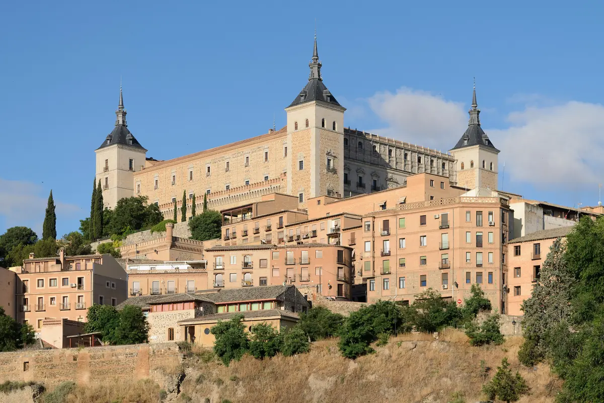 Alcázar v Toledu, rozsáhlá kamenná stavba s nárožními věžemi zakončenými tmavými špicemi, se tyčí nad domy s terakotovými střechami na úbočí kopce