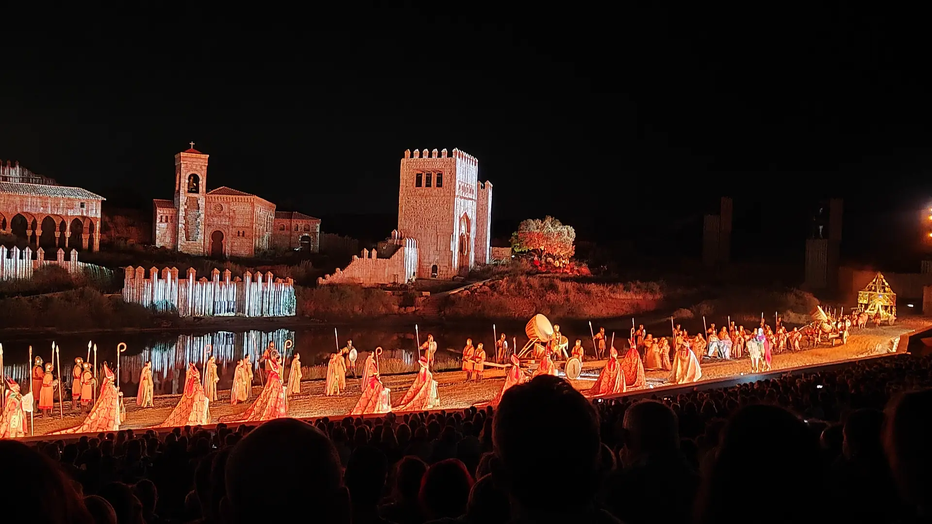 Noční show El Sueño de Toledo: dlouhý zástup účinkujících v oranžových a zlatých kostýmech kráčí po okraji jeviště před reflektory osvětlenými kamennými věžemi a kostelem, které se odrážejí ve vodě, zatímco v popředí jsou vidět hlavy diváků.